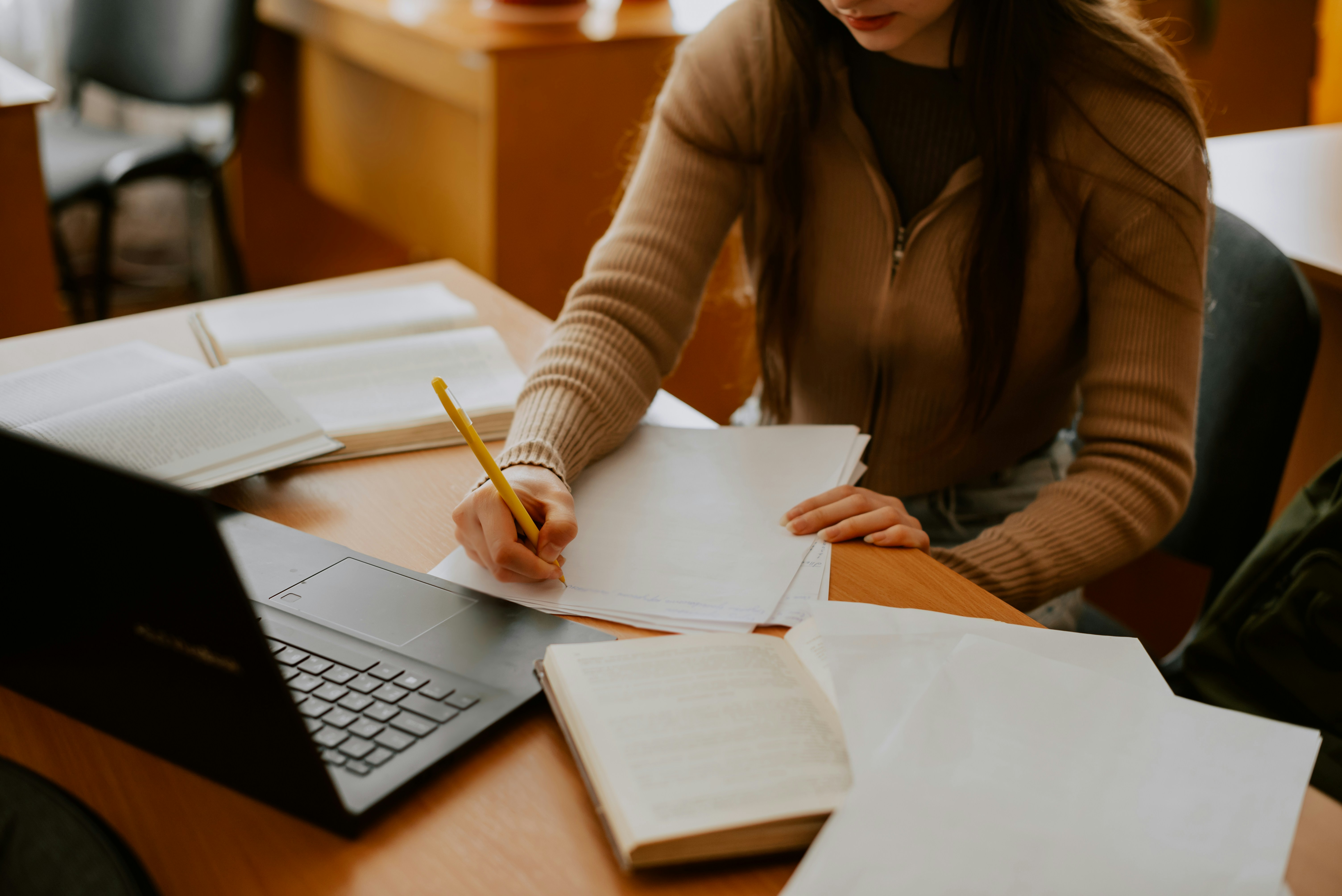 Person writing with a pencil at a table covered with papers and her laptop.