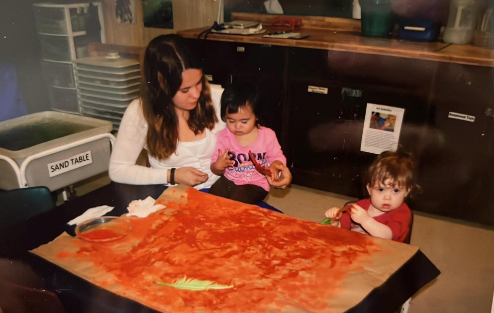 Woman holding a toddler while another toddler sits in a nearby chair. They're all painting at the table